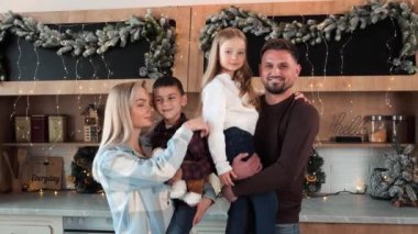 Dört kişilik mutlu bir aile. Erkek, kız, baba ve anne. Yeni yılı kutlamak için evde Noel çelenkleriyle süslenmiş mutfakta gülümsüyorlar. Evli çift çocuklarını kucağında tutuyor.