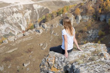Genç bir turist kız dağlarda bir uçurumun kenarında oturuyor ve doğanın güzel manzarasının tadını çıkarıyor..