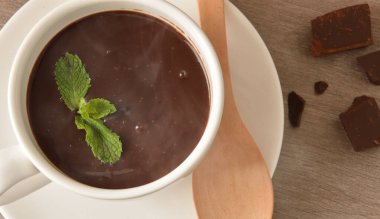 Tahta kaşık, nane yaprağı ve ahşap masa üzerinde çikolata parçaları olan beyaz seramik bardakta sıcak çikolata makrosu. Üst Manzara. Yatay bileşim.