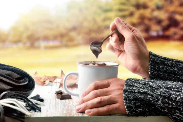 Cold hands with wool sweater stirring hot chocolate in a mug on wooden table with chocolate and wooden spoon with cinnamon and scarf and dry leaves with autumn landscape background. Front view