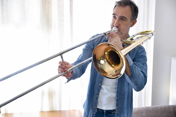 Trombonist practicing trombone in a room with curtain in the background at home.  Horizontal composition.