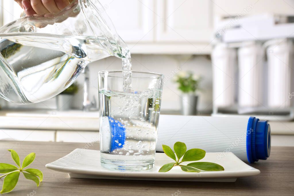 Hand filling crystal glass on kitchen bench from jug with filtered water using reverse osmosis household equipment. Front view. Horizontal composition.
