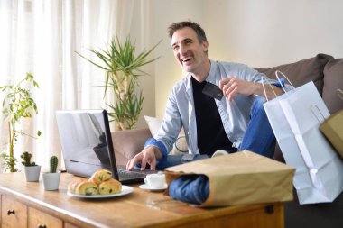 Neşeli adam evdeki kanepede dizüstü bilgisayarından alışveriş yapıyor. Yatay bileşim.