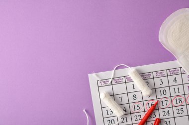 Regl takvimi geçmişi, işaretli kritik günler ve leylak arka planında çeşitli regl hijyen ürünleri. Üst görünüm.