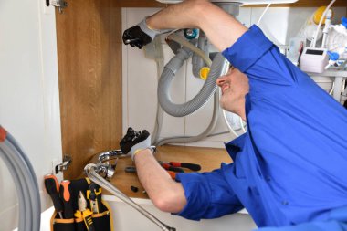 Üniformalı bir tesisatçı etrafta aletlerle lavabonun altındaki sızıntıyı tamir ediyor. Yatay bileşim.