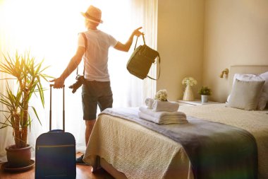 Bir otel odasında çantalı ve çantalı bir adam pencereden dışarı bakıyor. Güneş ışığı içeri giriyor.