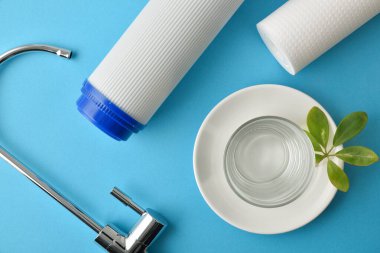Set of domestic reverse osmosis filters and glass with purified water on a plate with green leaves on a blue table. Top view. Horizontal composition.