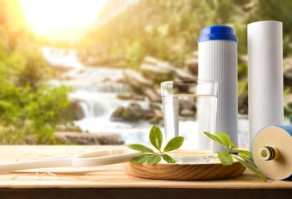 Glass of water purified by osmosis with environmentally friendly biodegradable filters on wooden slat table with nature background trees and waterfall. Front view. Horizontal composition.