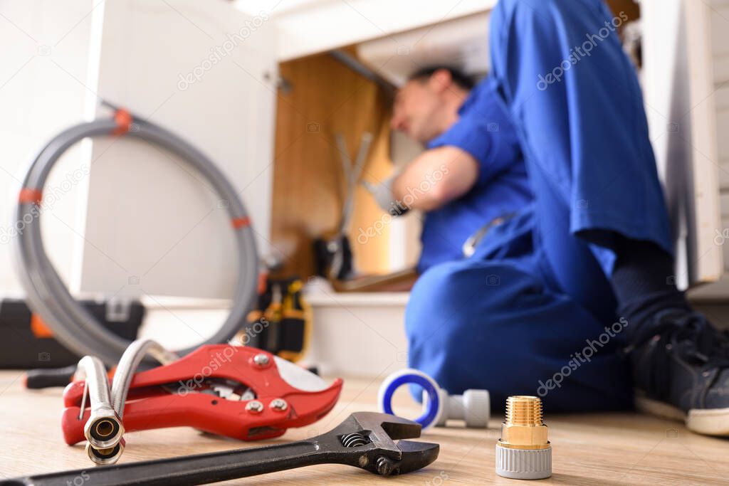 Plumbing material on a kitchen floor and a plumber doing a repair under a kitchen sink. Horizontal composition.