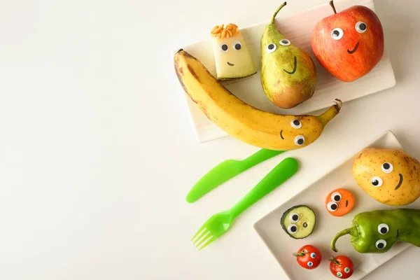 Background with plate of funny fruits with drawn eyes and mouths served for children on white table. Top view.