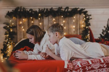Sevimli bir kız ve oğlan, Noel atmosferinde akıllı bir telefonla konuşuyor. Yeni yıl dekore edilmiş bir ev, çocuklar ve modern teknoloji, iletişim konsepti..