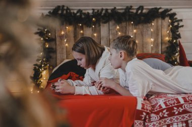 Sevimli bir kız ve oğlan, Noel atmosferinde akıllı bir telefonla konuşuyor. Yeni yıl dekore edilmiş bir ev, çocuklar ve modern teknoloji, iletişim konsepti..