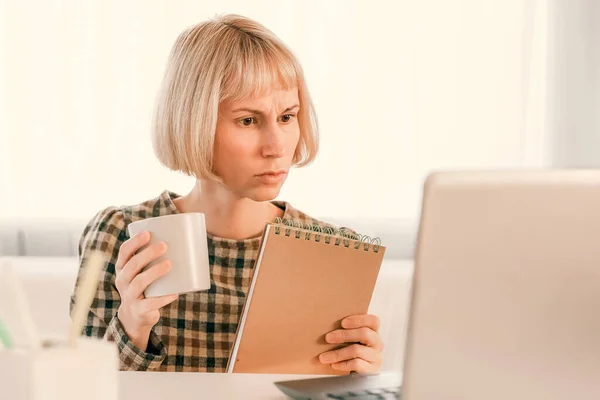 Woman watching on laptop display and writing notes. Video call, online conference, webinar