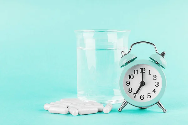 Alarm clock, a glass of water and white capsules with biological active additives or vitamins on a turquoise blue background. Selective focus. Copy space. Preventive and alternative medicine.
