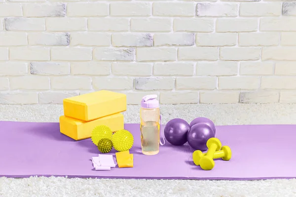 Fitness equipment on a yoga mat on a brick wall background. A bottle of water, rubber bands, yoga blocks, balls and dumbbells. Exercise equipment for home workout. Selective focus. Copy space.