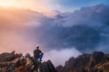 İnsan kayanın zirvesinde tek başına oturur. Yürüyüşçü ufukta sonbahar güneşini izliyor. Güzel bir an doğanın mucizesi. Vadideki renkli sis. Erkek yürüyüşü. Kişi silueti