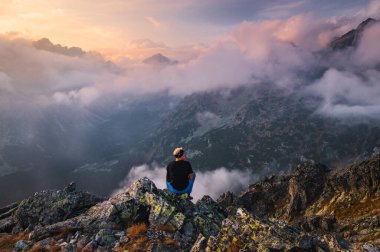 Siyah ceketli genç sakallı yürüyüşçü sis ve sisle çevrili dağlara bakıyor..