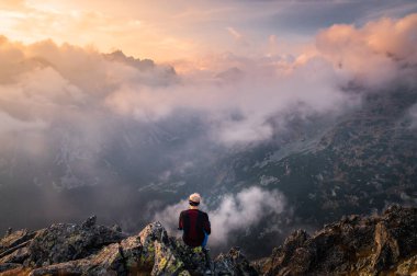 Kırmızı ceketli genç sakallı yürüyüşçü etrafı sis ve sisle çevrili dağlara bakıyor..