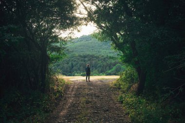 Female tourist walking on summer landscape on a trip in a green summer forest looking into the unknown at a yellow summer bow. Adventure photo in nature