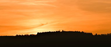 Gün batımında ufukta turuncu bulutlar görünüyor. Ormanla kaplı bir tepenin ardında..