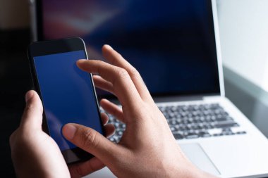 Model al, boş mavi ekran cep telefonu kullanan genç adam, ofis masasında dizüstü bilgisayarla dokunmatik ekrana yakınlaştırma, internet ödeme mobil uygulamaları veya web tasarım şablonu için kullan