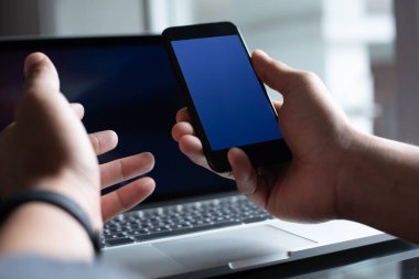 Boş mavi ekran cep telefonu kullanan, arka plan olarak dizüstü bilgisayarla dokunmatik ekrana yakınlaştıran, ürün görüntüleme, mobil uygulama veya web tasarım şablonu kullanan adam.