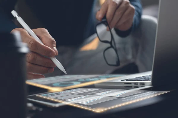 Business analysis and technology, financial investment concept. Business man reviewing marketing report on digital tablet and laptop computer with modern digital computer dashboard virtual screen in office, close up