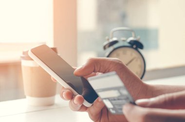 Adamın elleri cep telefonu ve kredi kartını ahşap masanın üzerinde sabah vakti evde bir fincan kahve ve saatle birlikte tutuyor. İnternet bankacılığı, online ticaret konsepti, ticaret konsepti, sıcak ton, klasik tarz..