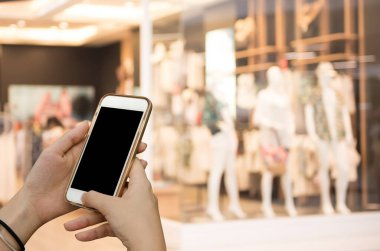 Çevrimiçi alışveriş, mobil bankacılık, internet ödeme konsepti. Alışveriş merkezinde arka planda bulanık moda mağazası olan boş ekran akıllı telefon kullanan genç bir kadının görüntüsü.