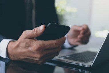 Business man in black suit using smart mobile phone and working on laptop computer, checking internet mobile apps in modern office, close up. Online working,  business and technology concept