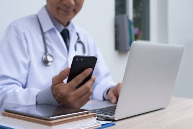 Smiling  30-40s asian doctor  looking at mobile smartphone and working on laptop computer with digital tablet on office desk , teleconference, e health service, health internet application on phone concept