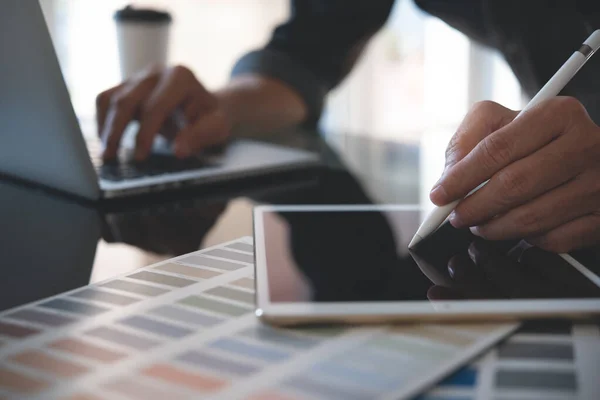 Website designer, graphic designer  creating project on mobile apps with stylus pen on digital tablet screen. Business man working on laptop and tablet computer with color swatch sample on desk in office, close up.