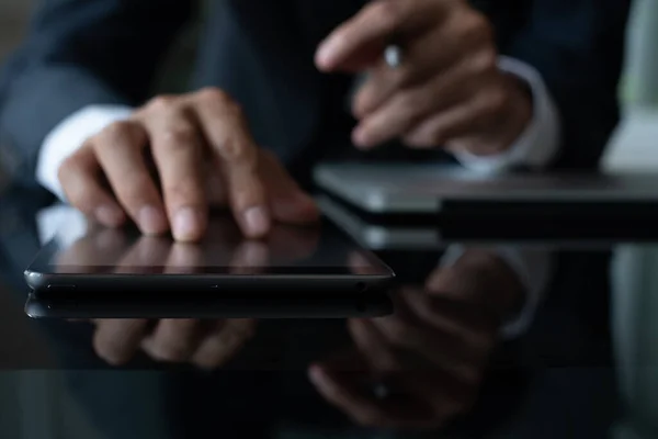 Businessman using digital tablet while working on laptop computer on desk with reflection, black tone, close up. Business and technology, Focus on work concept, Selective focus on tablet