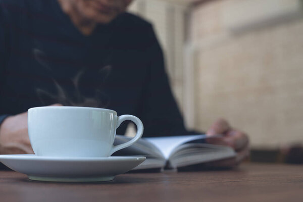 Closeup image of Asian man reading a book. Casual business man relaxed reading book with cup of hot coffee on wooden table in coffee shop or home office in the morning, vintage style.