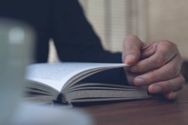 Closeup image of man reading a book. Casual business man relaxed reading book with cup of coffee on wooden table in coffee shop or home office, vintage style. Education concept