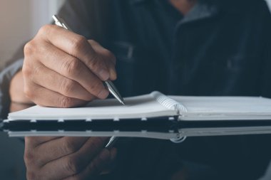 Man hand writing on papaer notebook with a pen. Man taking note or make a plan on notepad with reflection on office desk, close up, dark tone