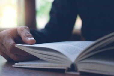 Closeup image of student reading a book in university library. Casual man opening and relaxed reading book on wooden table in coffee shop in morning sunlight, vintage tone.