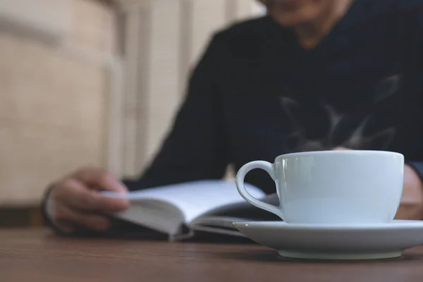 Closeup image of Asian man reading a book. Casual business man relaxed reading book with cup of hot coffee on wooden table in coffee shop or home office in the morning, vintage style.