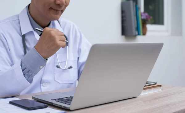 Portrait, smiling 30s 40s asian doctor working on laptop computer and looking at the screen in doctor room with smartphone on desk, telemedicine, EHRs Electronic Health Records system concept