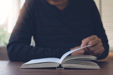 Closeup image of student reading a book in university library. Casual man opening and relaxed reading book on wooden table in coffee shop, vintage style.