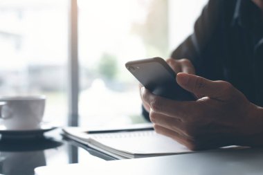 Casual man using smartphone with notebook and cup of coffee on desk at home, online working from home concept, close up