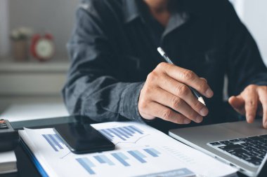 Buisness analysis concept. Businessman busy working on laptop computer, reviewing business data, financial report, mobile phone on desk in modern office with reflection and copy space, close up