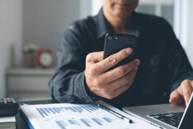 Asian business man  consultant using smart mobile phone analyzing  annual business report,  working on laptop computer with documents financial graph in office.