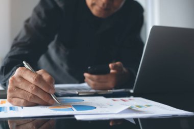 Buisness analysis concept. Businessman holding smart mobile phone busy working on laptop computer, reviewing business data, financial report on desk in modern office, close up