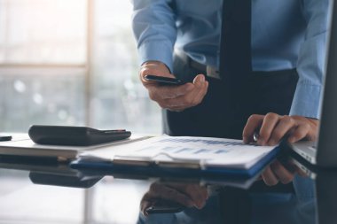 Business man or accountant using mobile smart phone, working on laptop computer with business document, financial graph diagram  on desk in modern office with sunlight. Business analysis concept, close up
