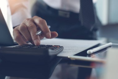 Close up of businessman or accountant hand holding pen working on calculator to calculate business data, accountancy document, laptop computer and digital tablet on  office desk, business concept. close up