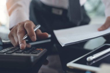 Close up of businessman or accountant hand holding pen working on calculator to calculate business data, accountancy document, laptop computer and digital tablet on  office desk, business concept. close up