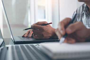 Two business men working on laptop computer, searching and taking note on paper notebook with pen. Students attending online class, Elearning, online working concept