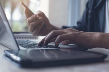 Cropped casual businessman, freelancer, blogger working on laptop computer, young man using internet browsing on digital tablet, mobile smart phone on office table with copy space, toned, close up