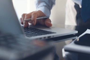 Work process concept. Business man typing on laptop computer keyboard with papaerwork and mobile smart phone on desk in modern office with blurry background, close up.